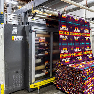 Fabric rolls with colorful geometric patterns exit a woolen mill machine in an industrial setting at Pendleton woolen mill washougal. Stacked fabric showcases vibrant hues.