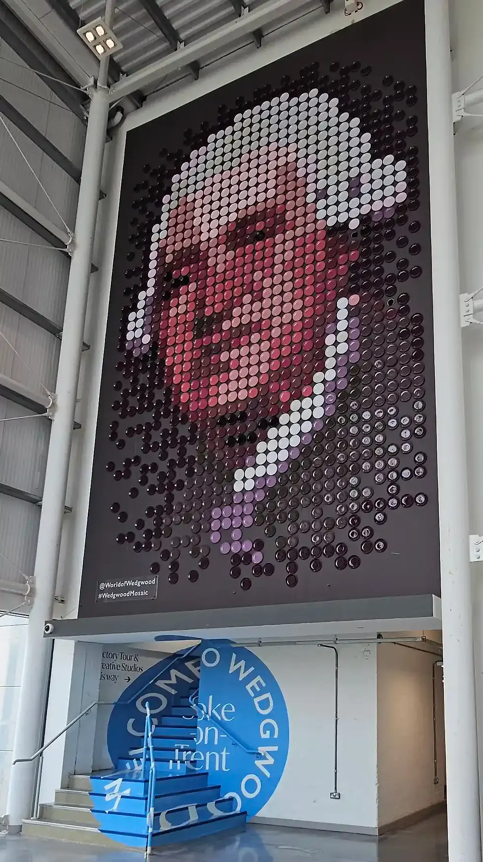 Mosaic portrait of Josiah Wedgwood in round plates on a wall, above blue staircase with "Welcome to Wedgwood" text. Bright lighting, industrial setting.