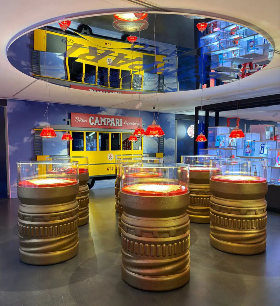 Golden barrel tables with red lighting, mirror ceiling reflections, yellow vintage tram backdrop with "Campari" text, vibrant decor. Image from Galleria Campari in Milan.