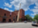 Red-brick building with giant baseball bat, American flag, and street with parked cars. Blue sky, fluffy clouds. People walking nearby.