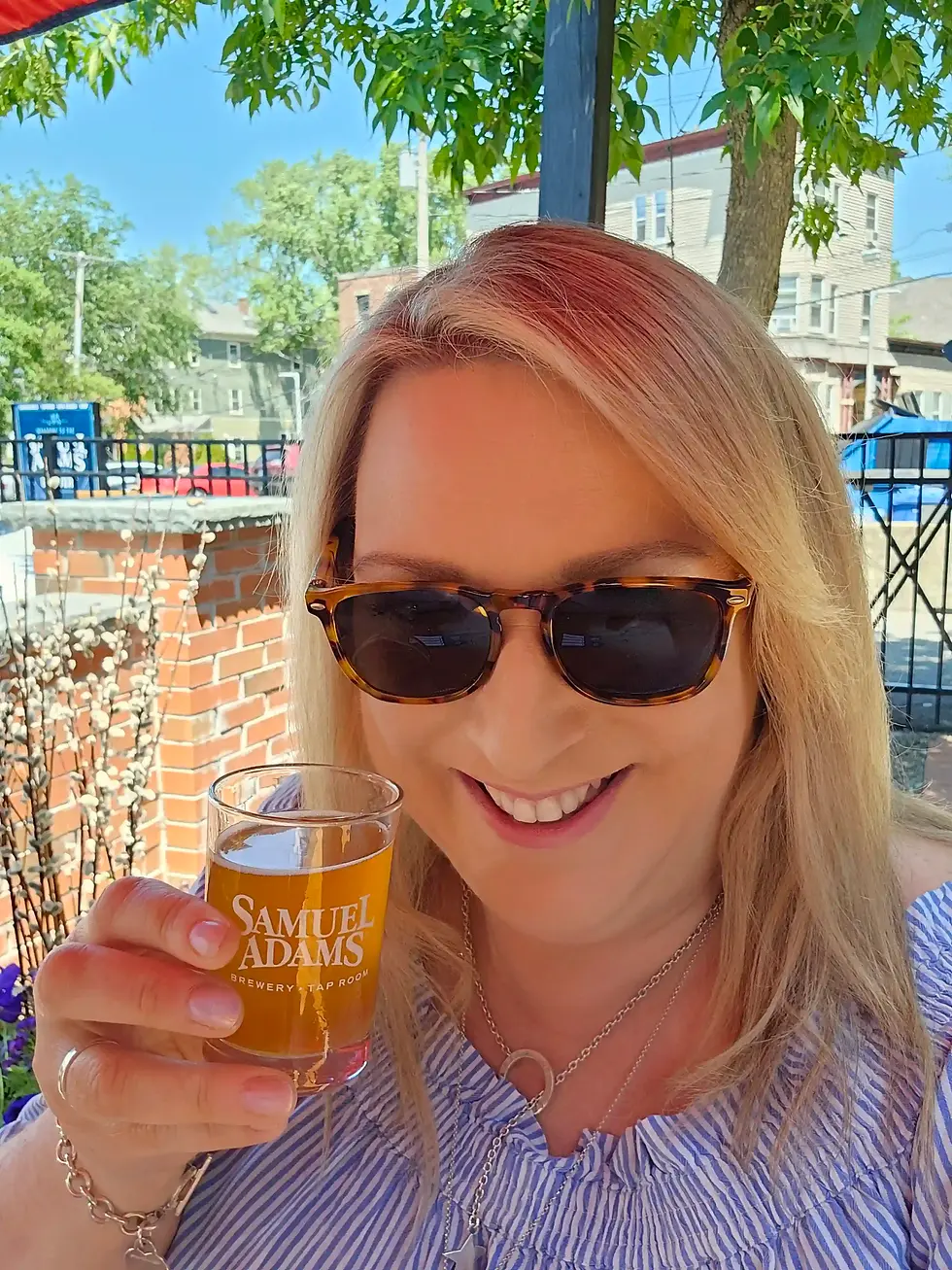 Smiling person in sunglasses holds Samuel Adams glass. Outdoor setting with trees, blue sky, and brick wall in background. Vibrant and cheerful mood.