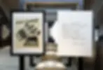 Framed Campari poster with fish and glasses design on left. Handwritten Italian text on white paper on right. Museum-like setting.