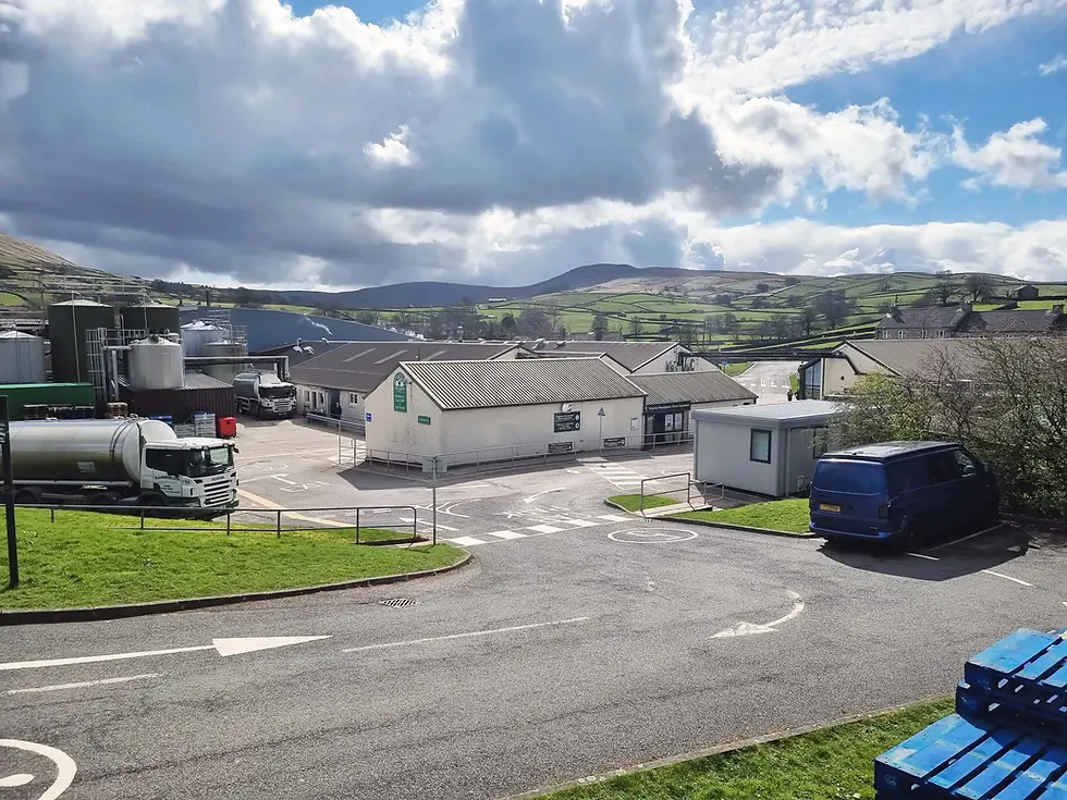 Trucks and buildings in an industrial area at wensleydale creamery under cloudy skies with distant green hills. A blue van is parked near blue pallets in the foreground.
