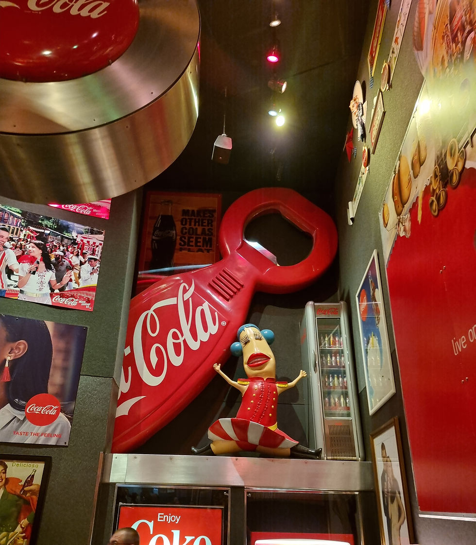 Coca-Cola themed room at World of Coca-Cola with vintage signs, a giant bottle opener, and a cartoon figure. People admire nostalgic displays under bright lights.