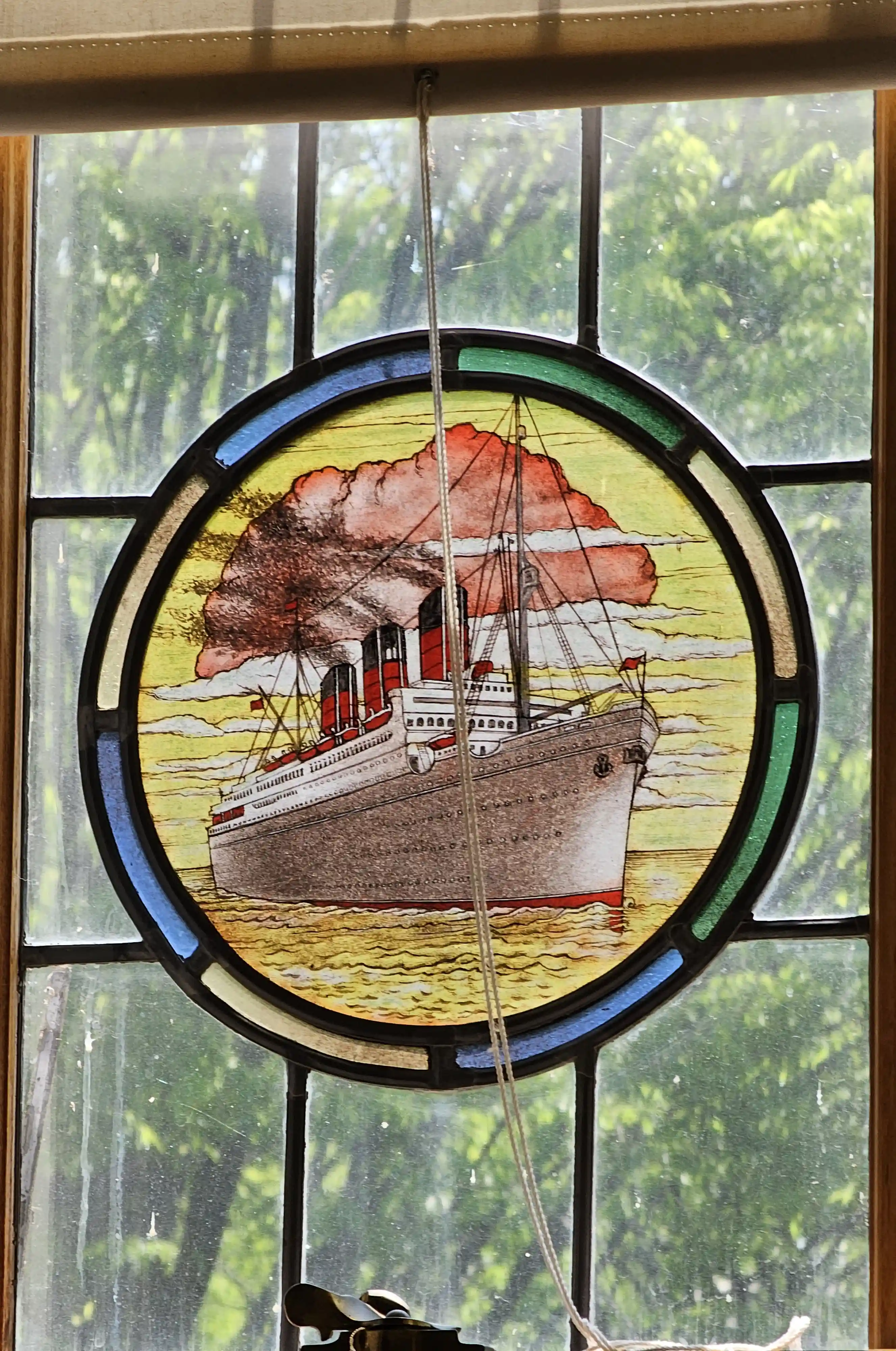 Circular stained-glass ship in a window, with green trees outside and a hanging blind cord, evoking a calm nautical scene at the George Eastman Museum.