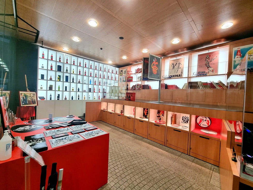 Shop display with a wall of colorful bottles, posters, and books inside Galleria Campari in Milan. A red counter holds flyers and a sanitizer bottle, under warm lighting.