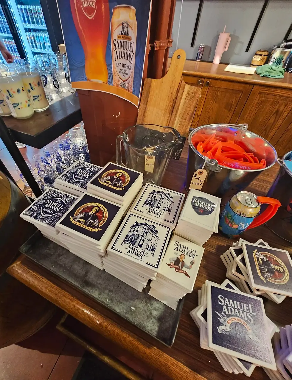 Samuel Adams Boston Brewery Tour shop display setup with Samuel Adams poster, stacks of themed coasters, and glassware. Colored cans and a bucket with orange scoops are visible.