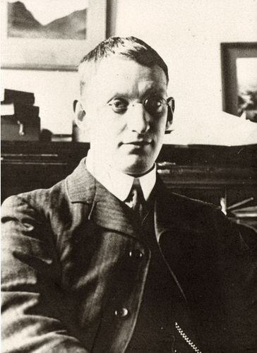 A man, Dr Kenneth Mees, in a suit and glasses sits in a dim room with framed pictures in the background. The image has a vintage black-and-white effect.