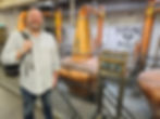 Man smiling with backpack in a whiskey distillery. Copper stills and "Teeling Whiskey" logo in background. Industrial setting, cheerful mood.