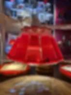 Red Campari Soda filled bottles hang upside down, forming a chandelier at the Galleria Campari. Background shows a colorful, blurred interior with art and books, creating a vibrant mood.