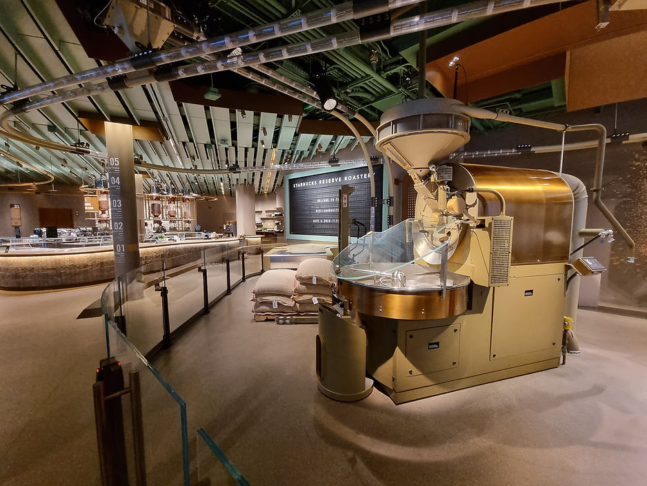 Starbucks Reserve interior with a large coffee roaster in the foreground, bags of beans, and a modern counter. Warm lighting and industrial decor.