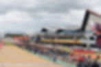 Crowds gather at Silverstone racetrack, with jet planes trailing red and blue smoke overhead. Lenovo signs and a large race building are visible.
