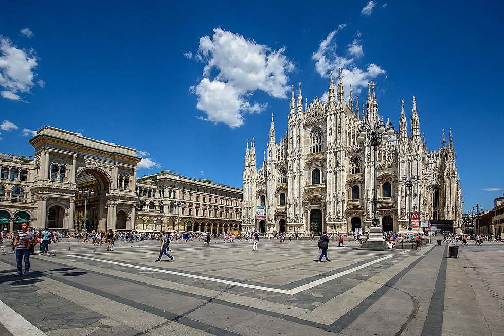 Crowd in sunny piazza with ornate white cathedral and surrounding historic buildings. Bright blue sky with fluffy clouds enhances vibrant scene.