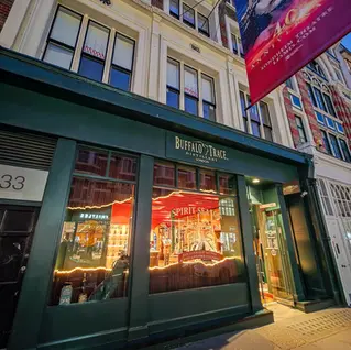 Street view of a building with a green storefront for Buffalo Trace Distillery London. A large red and blue banner hangs above, with neon lights inside.