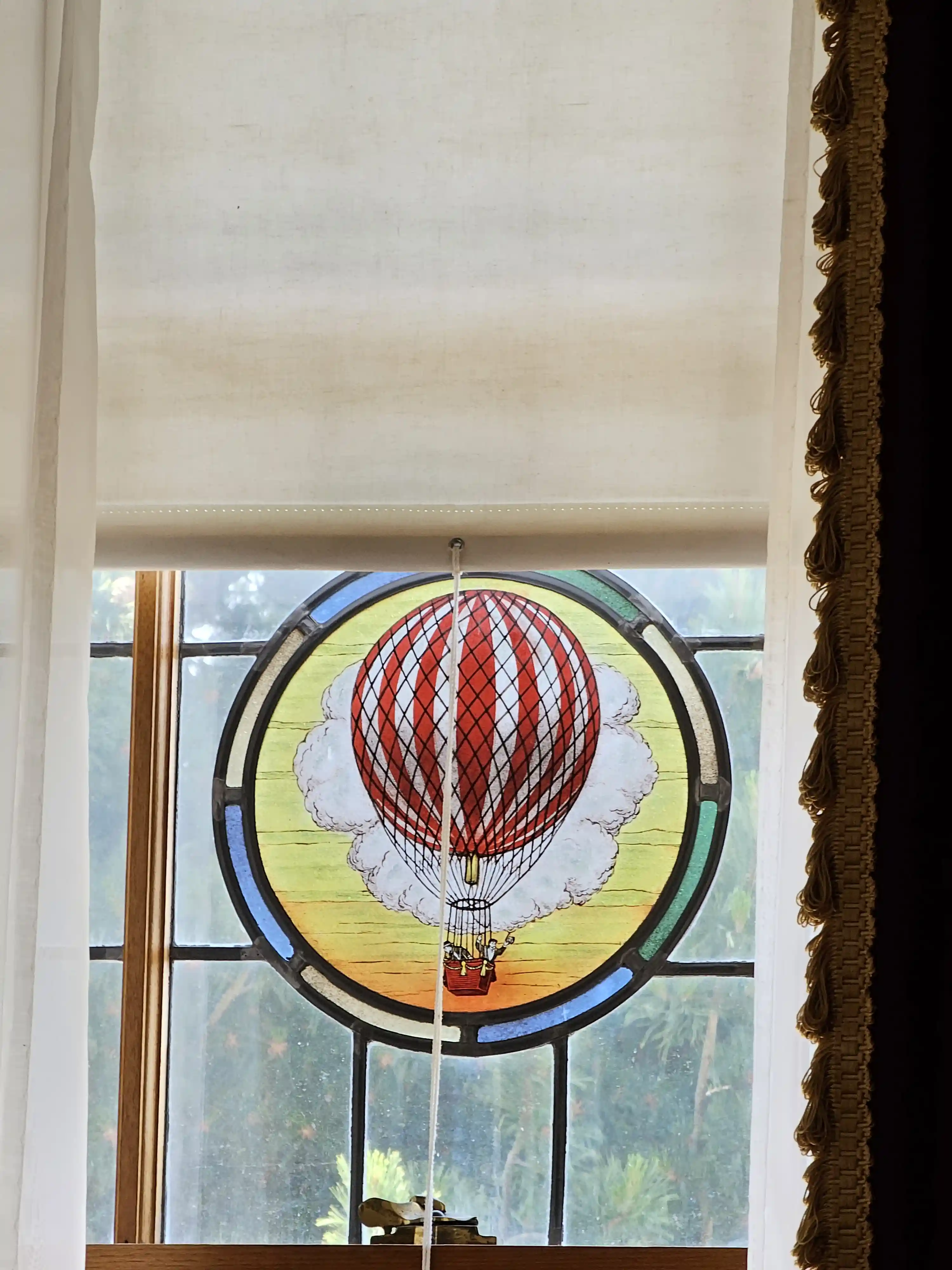 Stained-glass hot air balloon in a window, with white blind and tasselled curtain framing the colorful circular design at the George Eastman Museum.