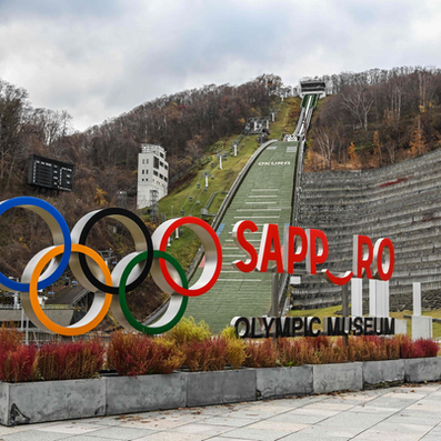 Olympic rings near ski jump at Sapporo, Japan. Overcast sky, autumn trees in background, "OKURA" written on ski slope.