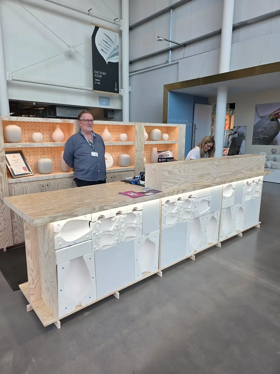 Man behind wooden counter with pottery display; woman working on computer. Modern interior with "Factory Tour" sign above at World of Wedgwood.