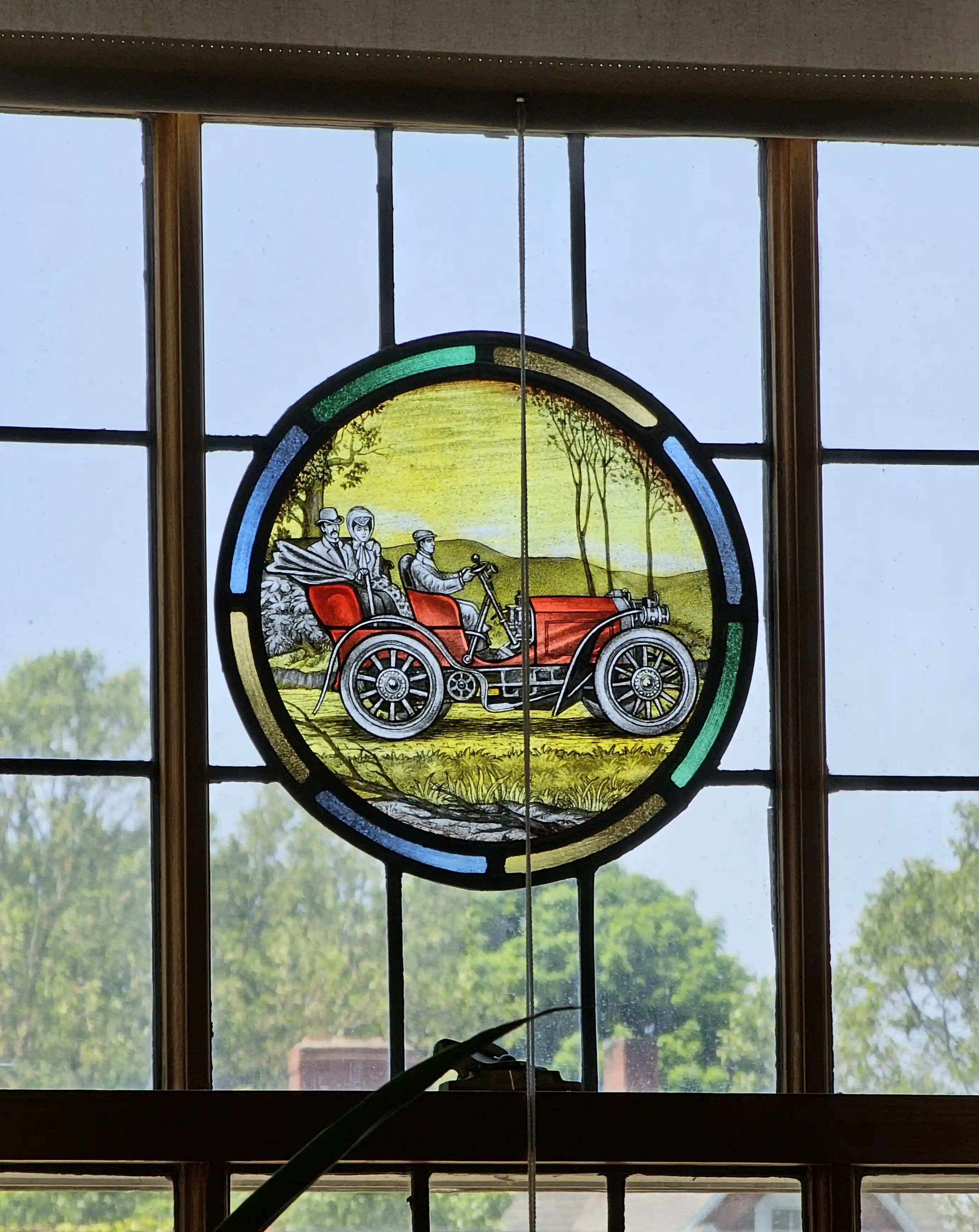 Round stained-glass window showing a red vintage car with three passengers on a sunny countryside scene at the George Eastman Museum.