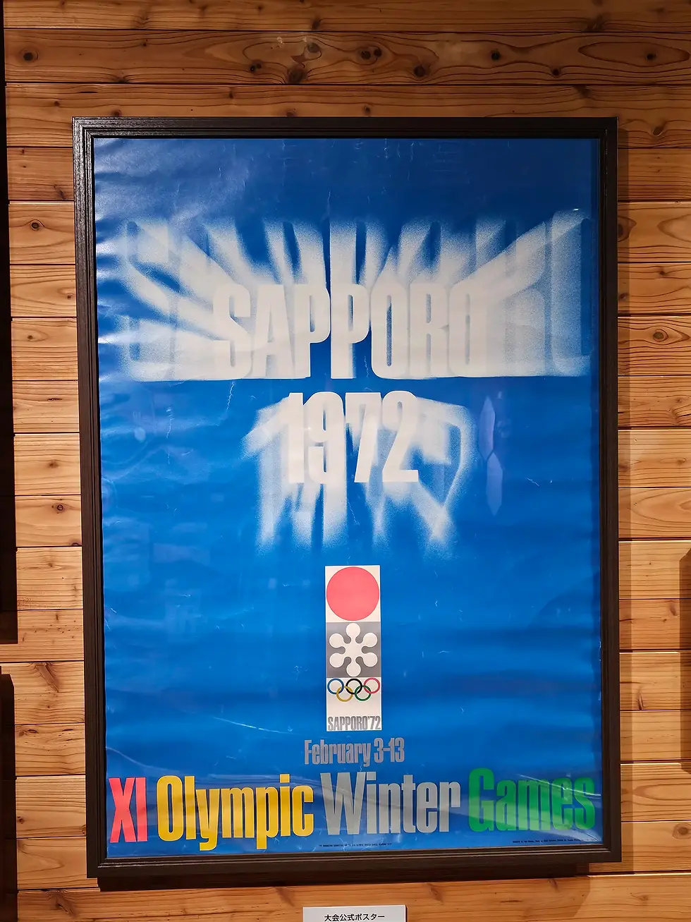 Blue poster for Sapporo 1972 Winter Olympics with bold text and Olympic rings. Wood-paneled background conveys a vintage vibe.