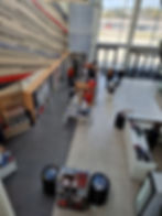 Silverstone museum shop interior with people browsing merchandise. Walls lined with shelves and colorful items. Bright daylight through large windows. Calm atmosphere.
