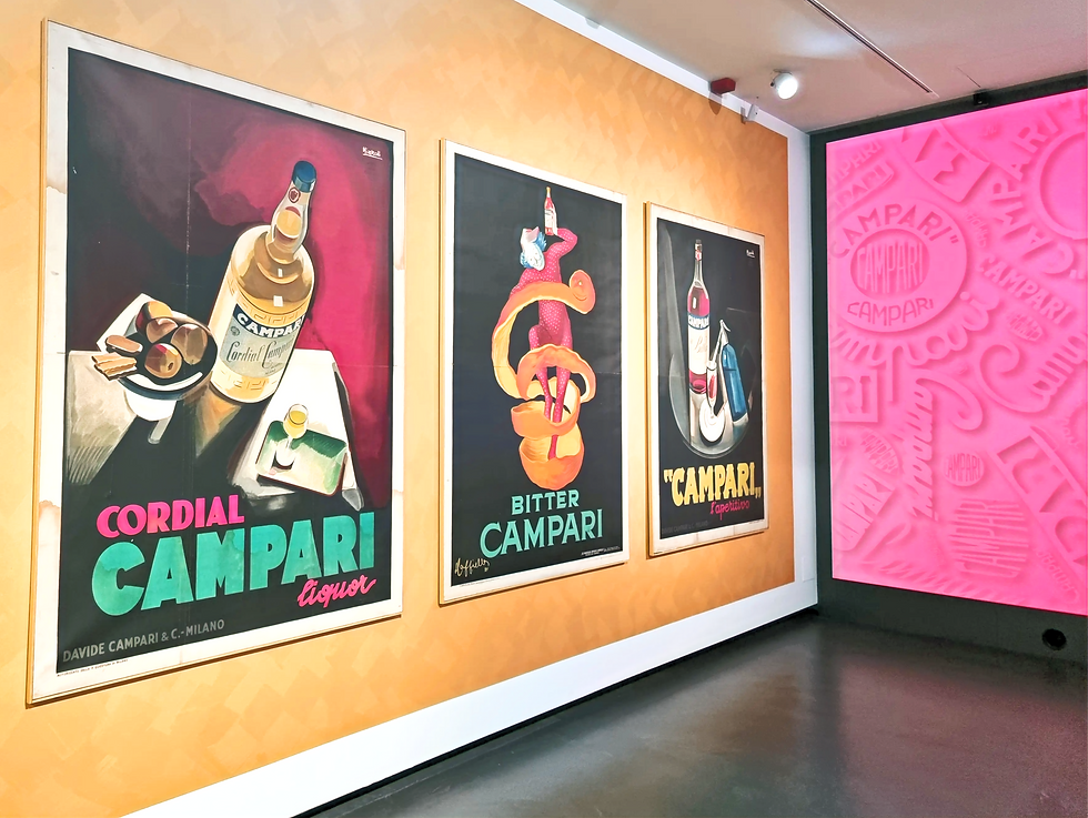 Three vintage Campari posters on a yellow wall, each featuring bold artwork and text. A pink wall with Campari embossed logos is on the right. Image from Galleria Campari in Milan.
