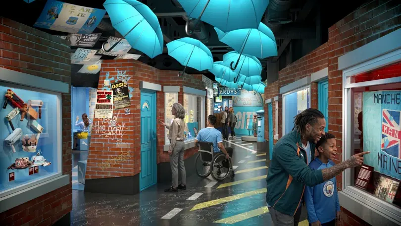 Visitors explore an indoor street exhibit of the planned new Manchester City Football club museum with bright blue umbrellas overhead, brick walls, posters, and a man in a wheelchair.