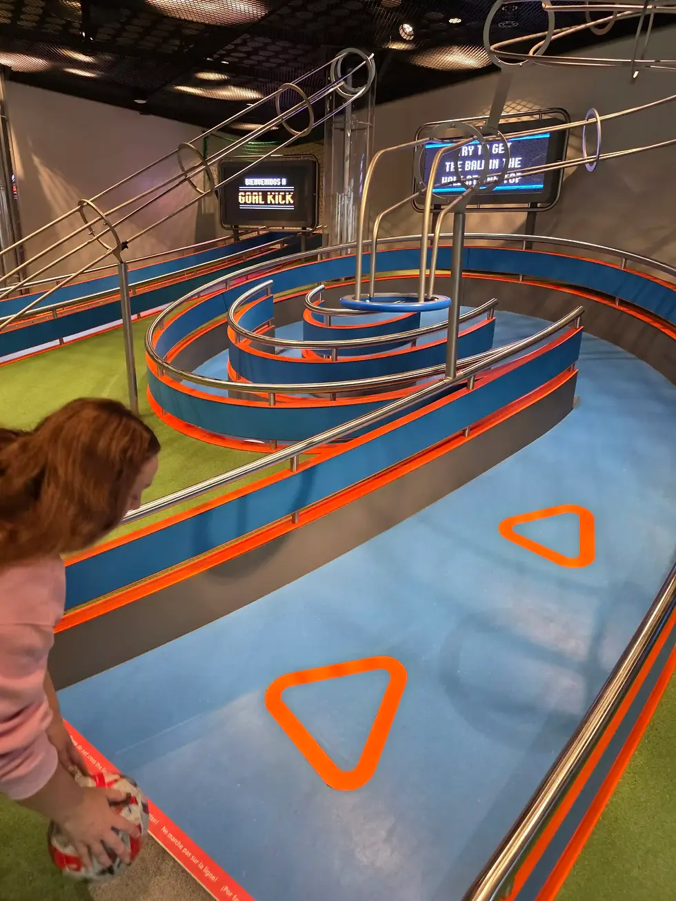 Child holding a ball beside a colorful indoor mini-golf course; screens read BIENVENIDOS A GOAL KICK at the FIFA Museum.