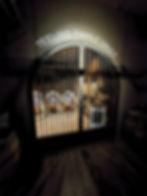 a darkened room has an arched opening in one wall with a metal gate hung in it, with metal bars all across and a large metal padlock on them. behind sits a small rack of barrels and 3 barrels on stands, and this room is lit