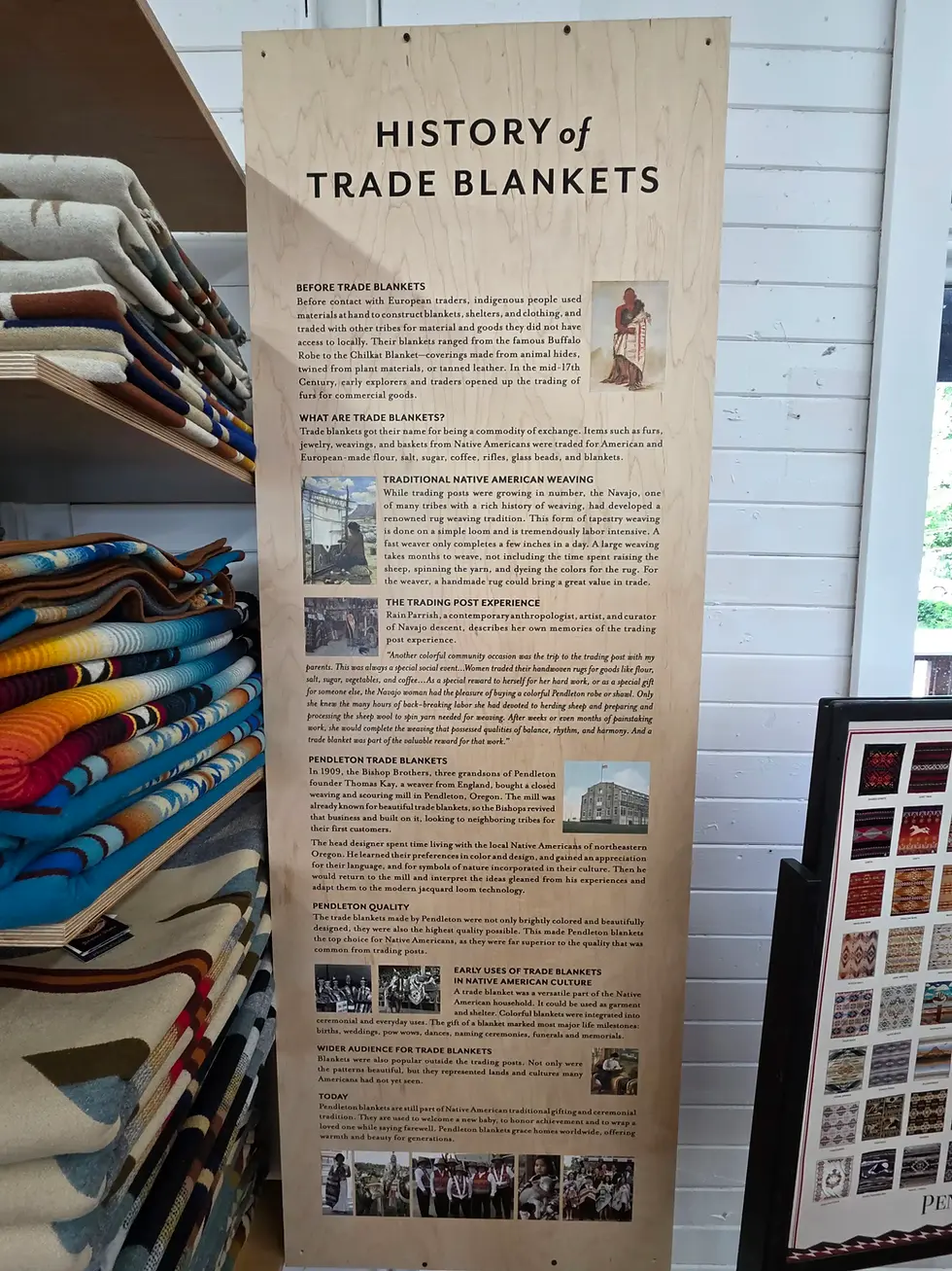 Display about the history of trade blankets with detailed text and images at Pendleton Woolen Mill Tour Washougal. Nearby, colorful folded blankets are stacked on a shelf.