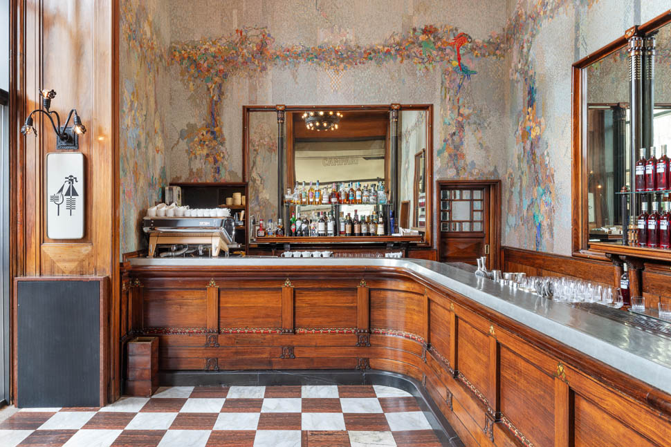 Elegant bar inside Camparino bar with wooden panels, mirrored back wall, and colorful mural. Bottles and glasses on the counter. Checkered floor adds charm.