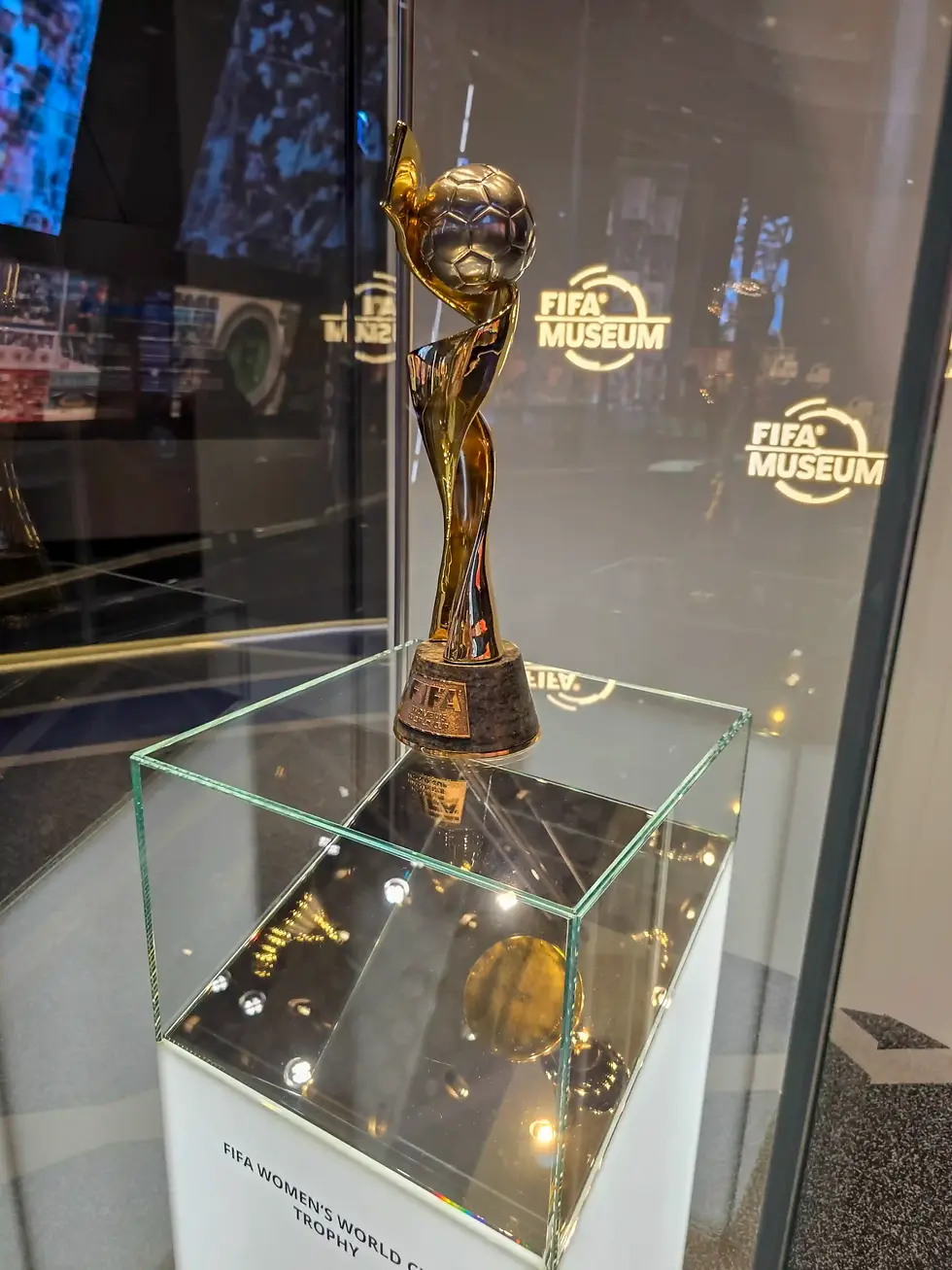 Golden FIFA Women's World Cup Trophy on a glass pedestal in a FIFA Museum display, with glowing logo reflections.