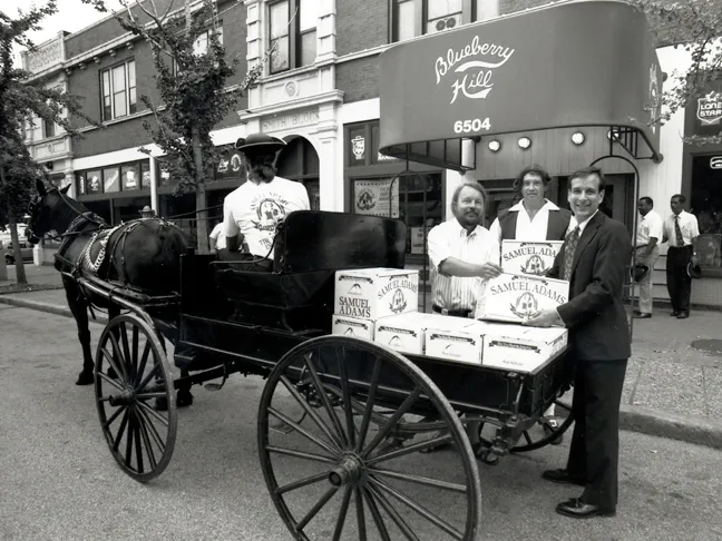 A horse-drawn cart with Samuel Adams boxes is parked in front of Blueberry Hill. Three men stand beside it, smiling, on a city street.