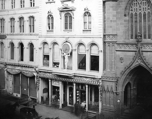 The Atlas Clock over the entrance to Tiffany & Co. 550 Broadway in the 1850s. Ornate historic building facade with arched windows and decorative details next to a gothic structure. Black and white photo.