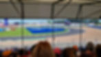 Race cars speed around Silverstone's Luffield corner, viewed from a covered grandstand with spectators. Blue and green colours dominate the scene.