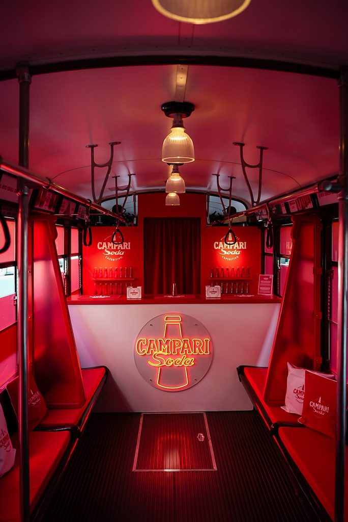Red-themed interior of a tram with Campari Soda branding, featuring a bar, illuminated logo, seating, and hanging lamps, creating a vibrant ambiance.