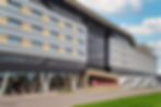 Modern hotel exterior with large windows, geometric patterns, and "Hilton Garden Inn" sign. Mostly gray facade under a partly cloudy sky.