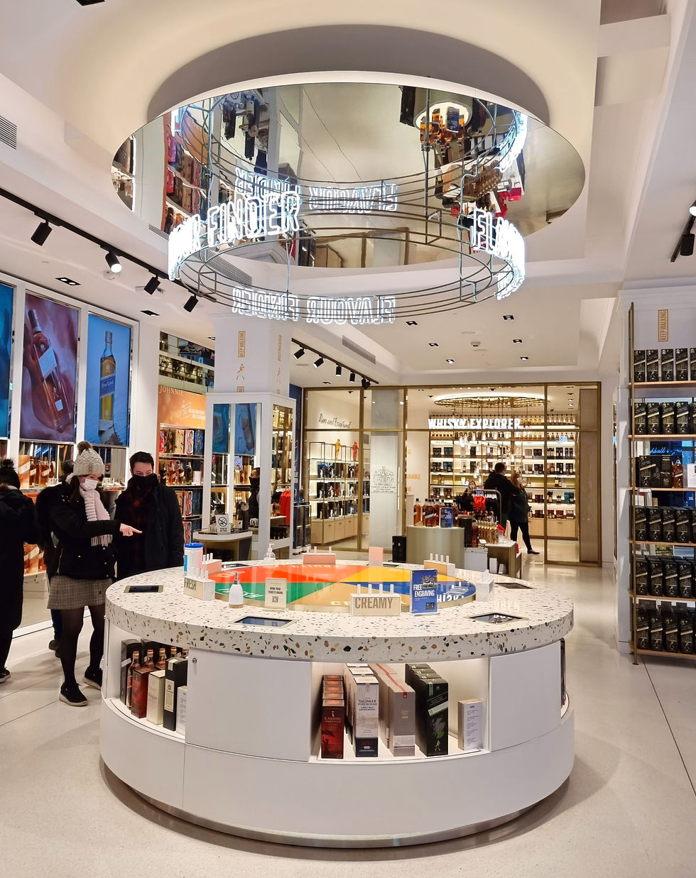 Shoppers in a brightly lit store explore whiskey displays. Neon sign reads "FLAVOUR FINDER." Circular counter with colorful sections.