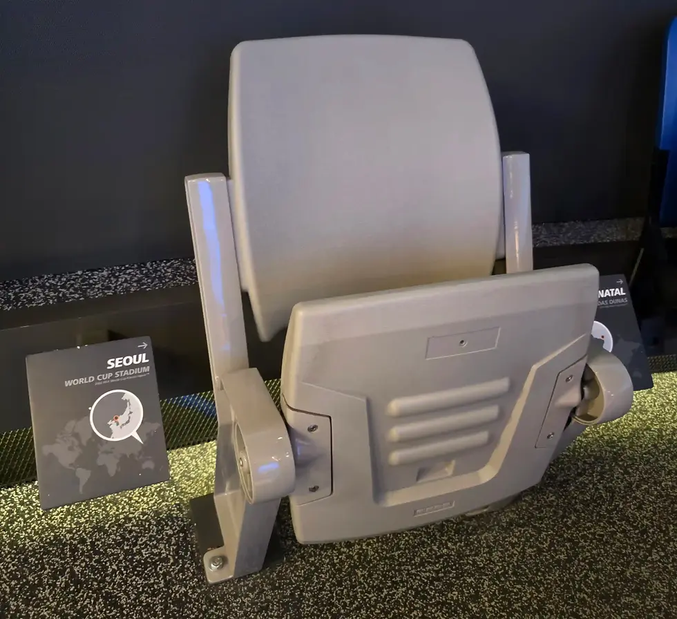 Gray stadium seat display in a museum, with signs for Seoul World Cup Stadium and Natal Das Dunas at FIFA Museum.
