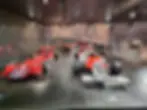 Red and white vintage racing cars displayed in a dimly lit museum with driver portraits in the background. Text includes "Marlboro" and "Martini."