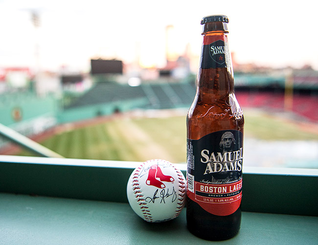 A Samuel Adams Boston Lager bottle and a baseball with the Red Sox logo sit on a railing at a baseball stadium, with the field blurred behind.