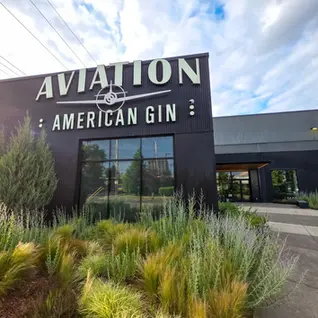 Black building with "Aviation American Gin" sign, surrounded by lush greenery. Sunny day with a blue sky and clouds.