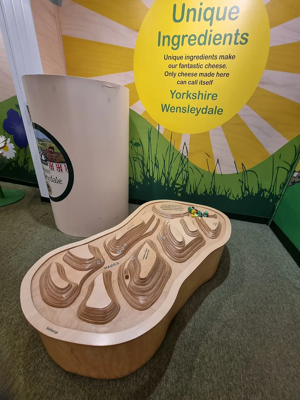 Wooden topographical model with labeled hills and small toy figures. Yellow sign reads Unique Ingredients, Yorkshire Wensleydale. Green setting.