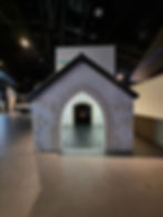 Small grey house structure in a dimly lit room in the Silverstone museum, with a passage leading into a dark interior. "Fire exit" sign on the wall above.