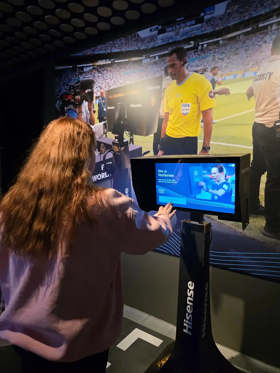 Woman uses a Hisense touchscreen to try a FIFA VAR referee game in front of a stadium backdrop at the FIFA Museum.