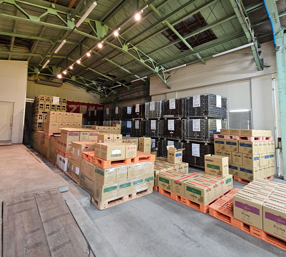 Warehouse interior with stacked cardboard boxes on orange pallets and metal crates under green beams, some Japanese signs visible as seen on the Lumiere winery tour.