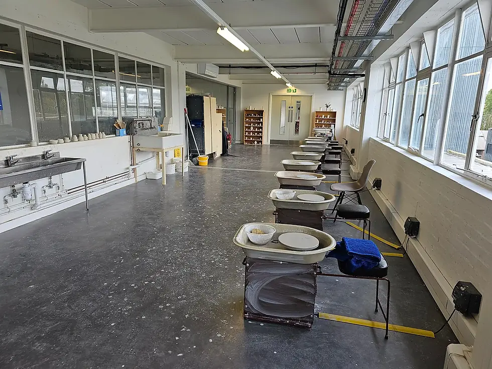 Empty pottery studio at World of Wedgwood with several pottery wheels and chairs lined up. Bright space with large windows, gray floor, and sinks on left.