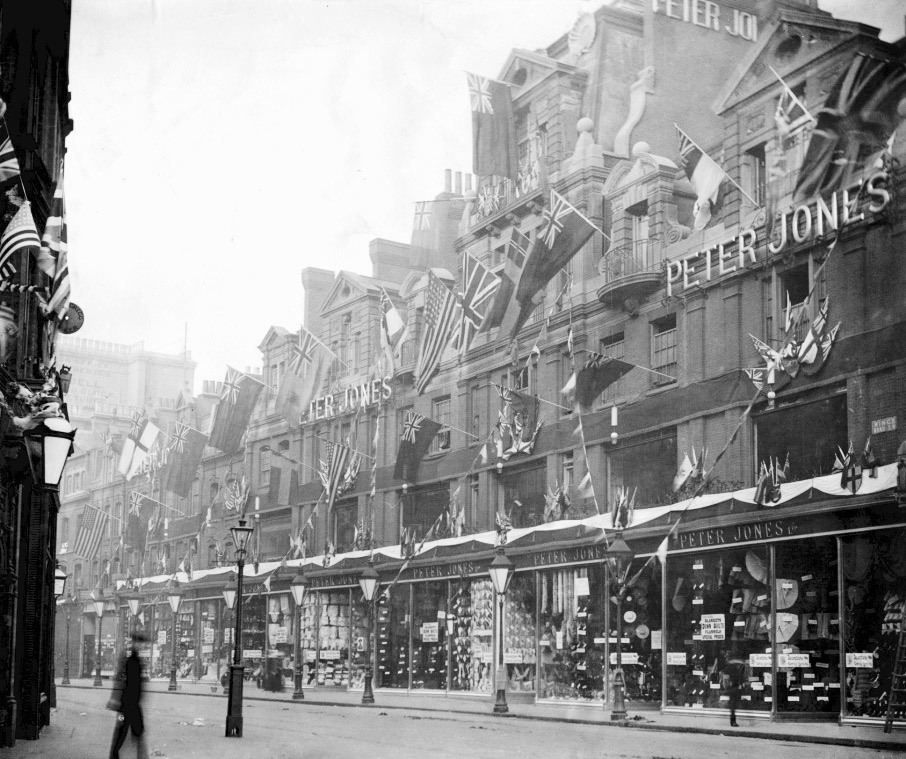 Street scene with a row of Peter Jones storefronts, decorated with numerous flags. A person walks by. Historical, busy atmosphere.