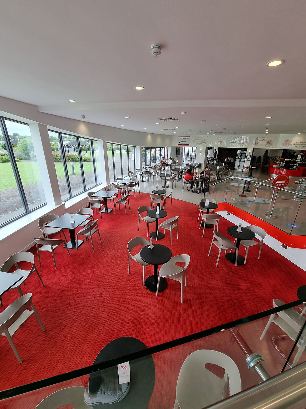 Spacious cafe at the Haynes Motor Museum with red carpet, black tables, and beige chairs. Few people seated. Large windows show greenery outside. Calm atmosphere.