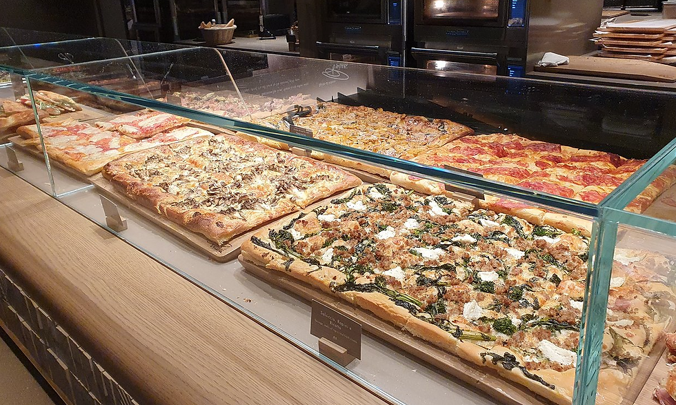 Rectangular pizzas with various toppings displayed in a glass case on a wooden counter. Background shows ovens in a warm-lit setting.