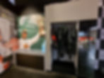 Silverstone museum dressing up area with racing overalls hanging beside a poster of Lewis Hamilton holding a trophy labeled "2017 Rolex British GP, Silverstone." Cars and checkered pattern in background.
